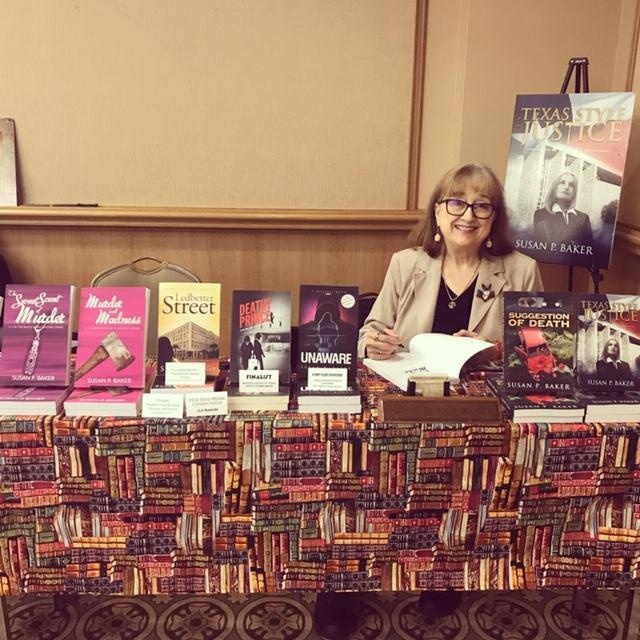 The author Susan P. Baker is seated behind a table covered with a tablecloth with a pattern of book spines arranged in rows. Her own books are displayed on top of the tablecloth. Her body is seen from bust up in a gap between the books displayed. She is wearing a black shirt with a tan collared jacket over it and a necklace. On her jacket is a circular brooch. She is smiling and holding a book open against the table with a pen, as if she is going to sign it. Behind her is a tan wall with a wood paneling that rises midway up the wall. Behind her and to her left, in the right side of the picture, a promotional display with the cover of her novel Texas Style Justice is resting on a display easel.