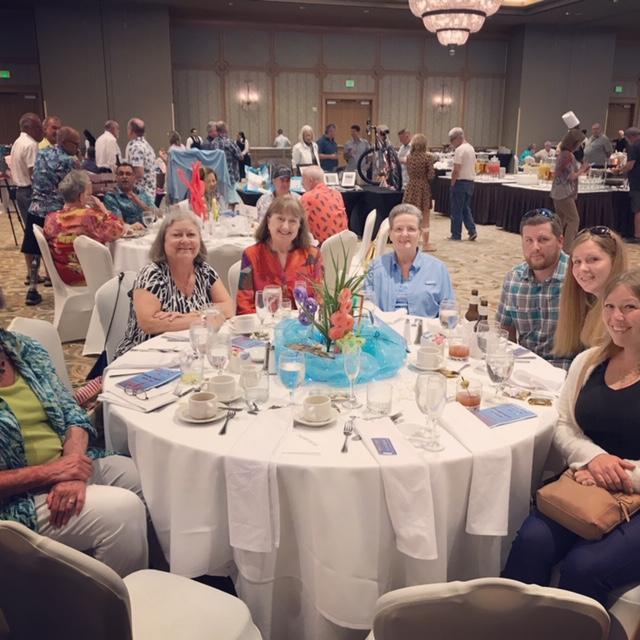 The author Susan P. Baker is pictured at a fundraising event in a large ballroom. She is seated close to the middle of the image at the far side of a large round table with a white tablecloth, a tropical centerpiecem and numerous used dishes and glasses. There are 6 other people seated around the table with her, and all are facing the camera and smiling. Ms. Baker herself is wearing a reddish orange top with long patterned sleeves. Beyond the table with Ms. Baker and her companions, there are a large amount of people sitting at other tables or standing and walking throughout the ballroom. At the top of the image is the bottom of a large glass chandelier and it is reflected in the glass windows at the far back of the ballroom.