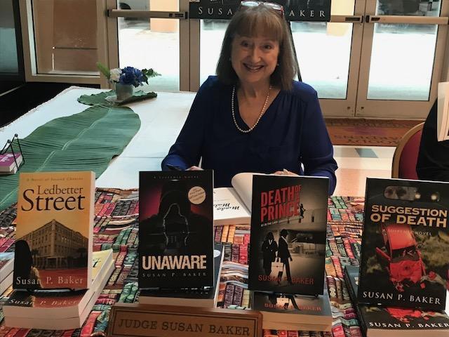 The author Susan P. Baker sits in a chair behind a table where several of her books are displayed on top of a tablecloth that has a pattern of book spines lined up in rows. Behind her are glass doors and to her left is another table with a green table runner and other unidentifiable objects on it. Ms. Baker herself is wearing a navy blue long sleeved top, a long white necklace, and her glasses are on top of her head. She is smiling at the camera and holding open a book against the table as if she is going to sign it.
